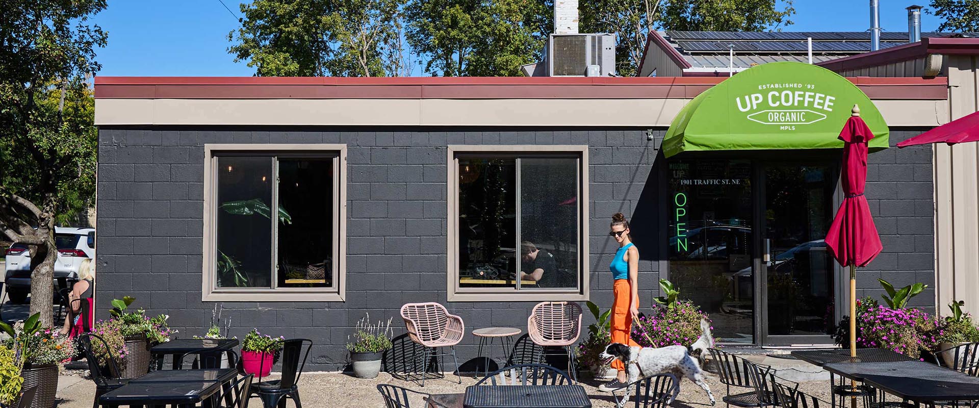 Outdoor patio area of a coffee shop with black walls, large windows, and a green awning displaying the text 'UP COFFEE ORGANIC MPLS.' A woman in bright clothing walks a dog past colorful chairs, tables, and flower pots under a sunny sky.