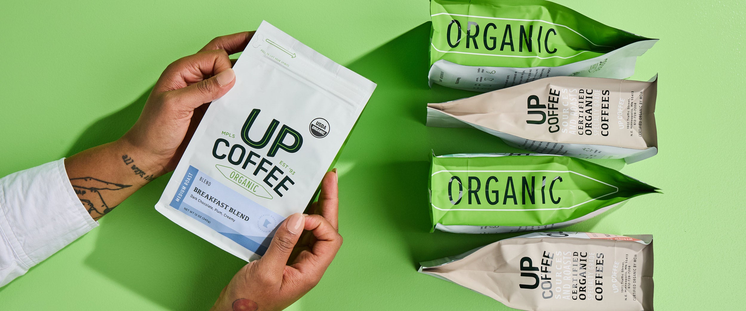 A person with a tattooed arm wearing a white sleeve holds a white bag of "UP Coffee Organic" Breakfast Blend, a medium roast coffee. The packaging features a USDA Organic label. On a green background, four additional coffee bags with green and beige packaging, labeled "ORGANIC" and "UP Coffee," are arranged in a row.
