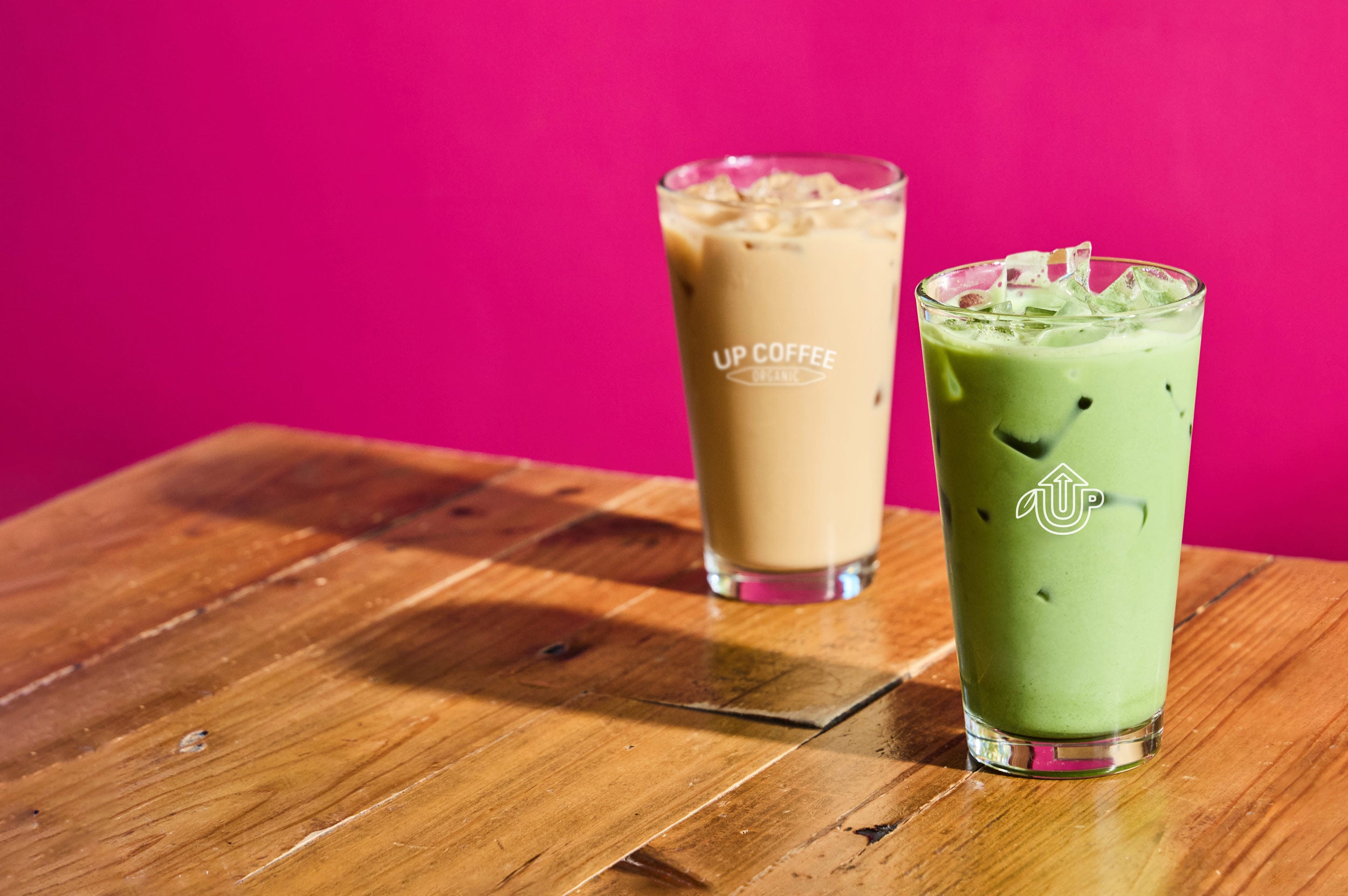 Two iced beverages in tall glasses sit on a rustic wooden table against a vibrant pink background. The front glass contains a green matcha drink with ice, featuring the "UP Coffee" logo. Behind it, a creamy iced coffee with ice bears the "UP Coffee Organic" branding. The drinks cast soft shadows on the table.