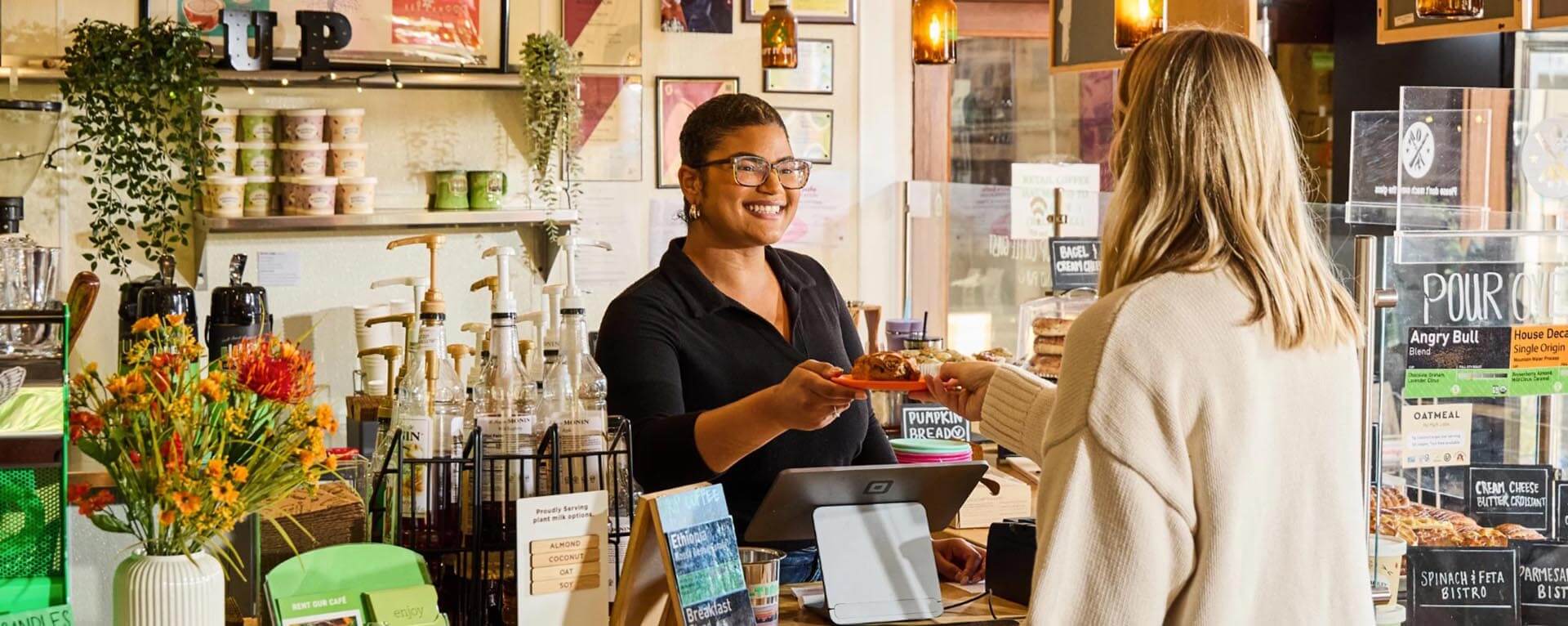 A friendly barista in a cozy coffee shop hands a pastry to a customer across the counter. The counter is adorned with syrups, menu items, and vibrant flower arrangements.