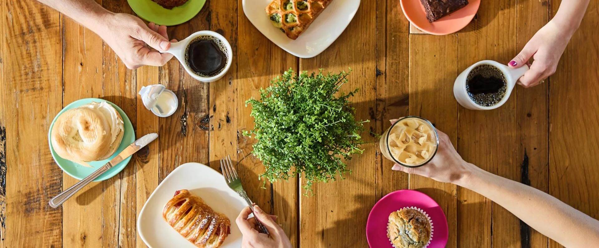 Top-down view of a wooden table set with various pastries, coffee cups, and an iced coffee, surrounded by vibrant plates and a small potted plant in the center.